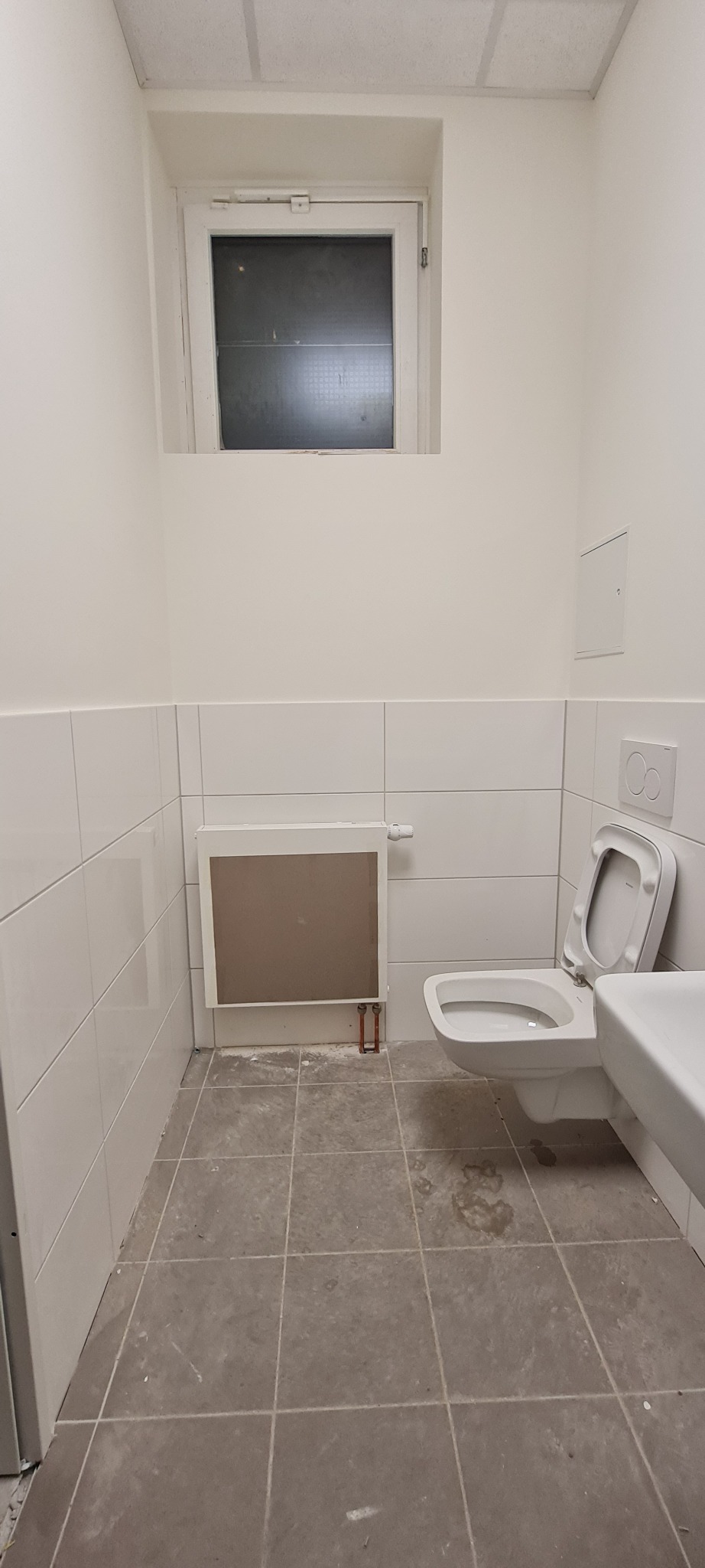 Wnętrze łazienki po remoncie: białe płytki na ścianach, szare płytki podłogowe, grzejnik panelowy, toaleta z otwartą klapą i umywalka, małe okno pod sufitem, widoczne ślady po remoncie na podłodze.