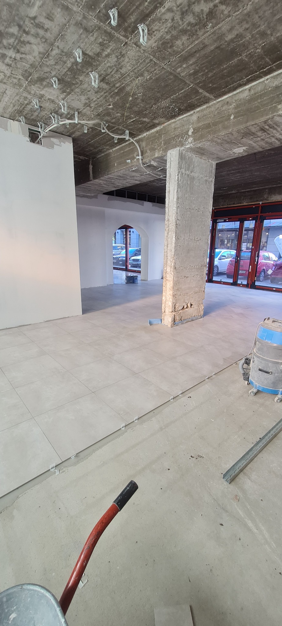 Wnętrze w trakcie remontu z ułożonymi płytkami podłogowymi, widoczny surowy betonowy sufit z instalacją elektryczną, kolumna w stanie surowym, odkurzacz przemysłowy, taczka i profile metalowe.