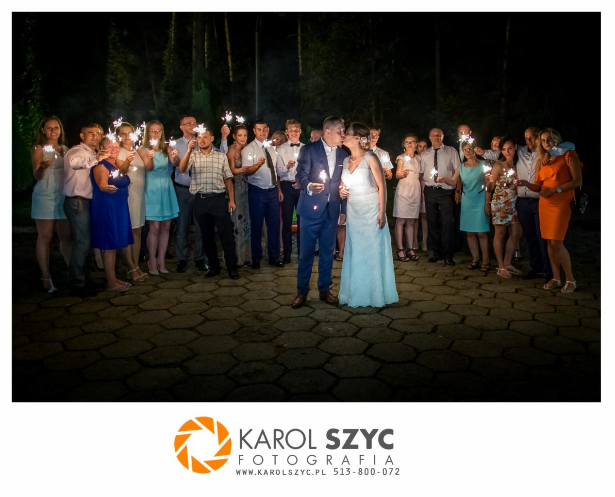 Pocałunek nowożeńców na tle gości weselnych trzymających zimne ognie, wieczorny plener.