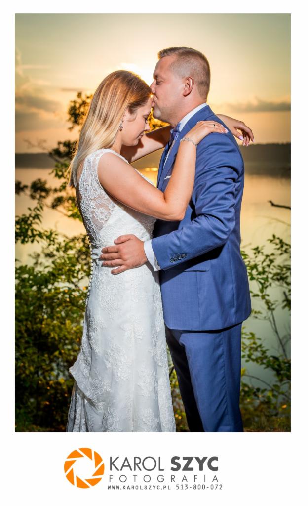 Para młoda w objęciach na tle jeziora o zachodzie słońca; pan młody w niebieskim garniturze całuje pannę młodą w koronkową suknię.
