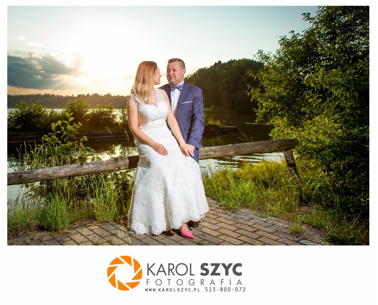 Para młoda w plenerze nad jeziorem, panna młoda w koronkowej sukni z różowymi butami, pan młody w granatowym garniturze z muszką, drewniana barierka w tle o zachodzie słońca.