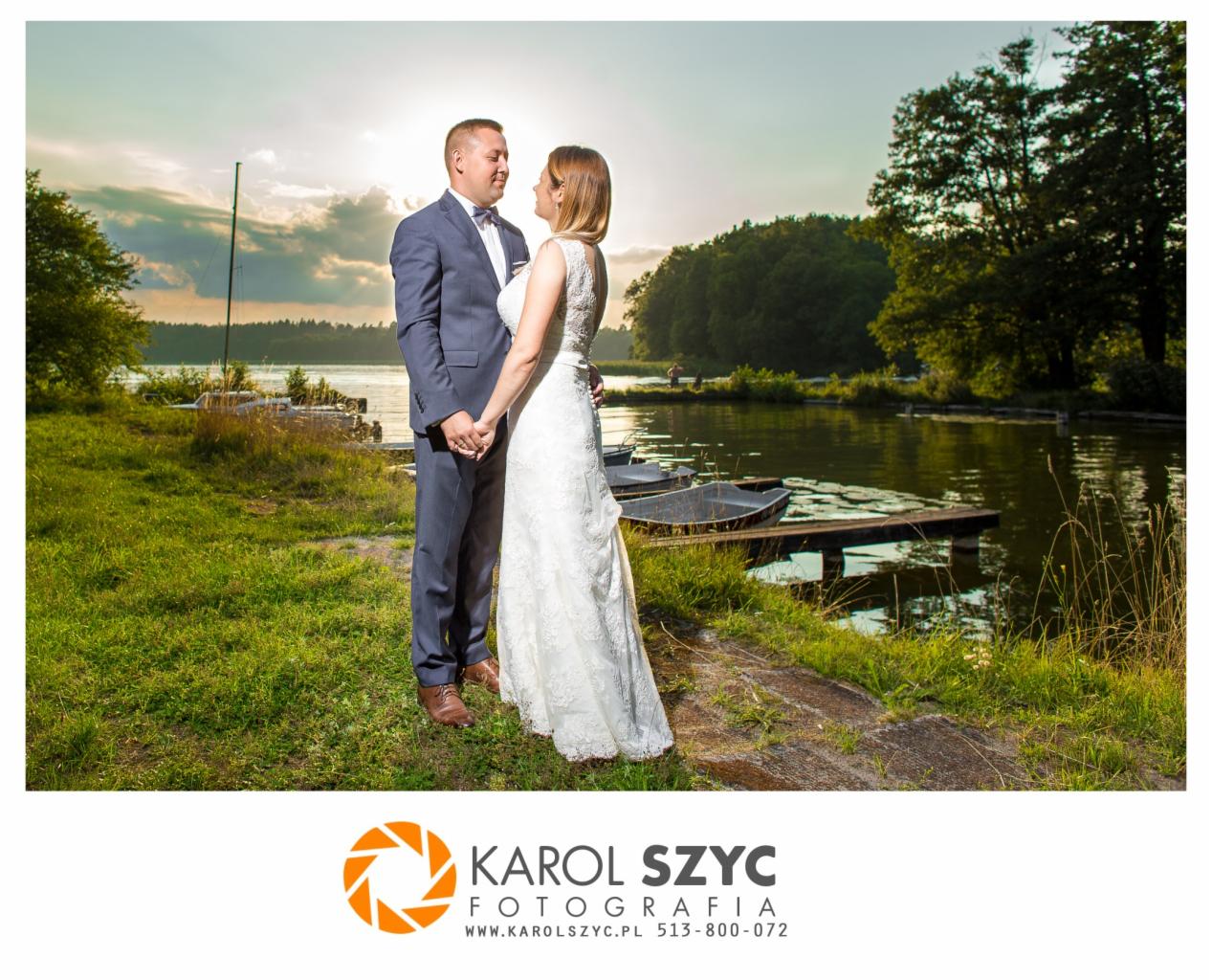 Para młoda trzymająca się za ręce, pozująca na tle jeziora i zachodzącego słońca, łódki na brzegu, mężczyzna w granatowym garniturze, kobieta w białej koronkowej sukni ślubnej z odkrytymi plecami.