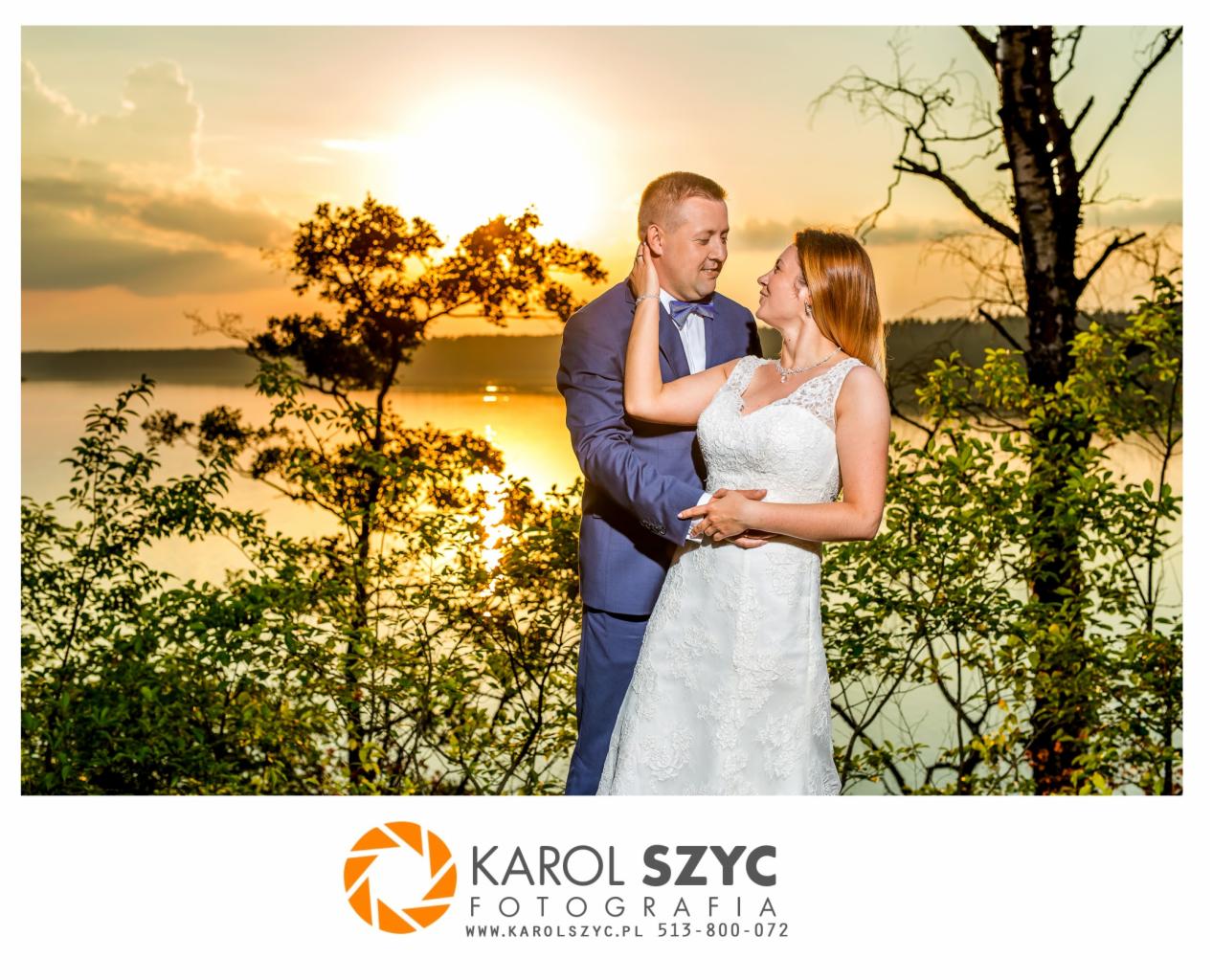 Para młoda w objęciach na tle jeziora o zachodzie słońca, on w niebieskim garniturze, ona w białej sukni ślubnej z koronkową górą, drzewa i krzewy w tle.