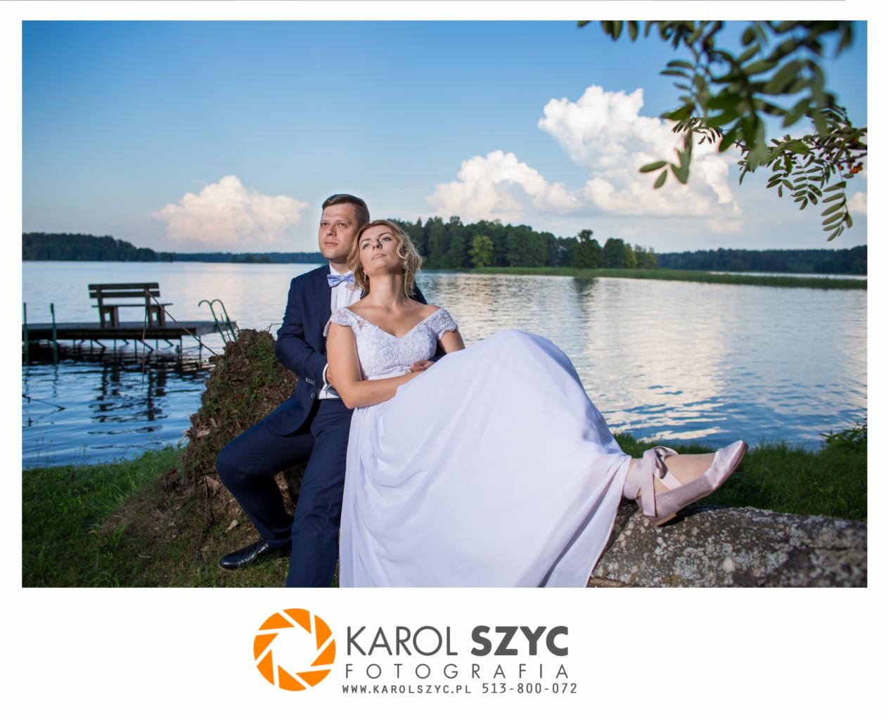 Para młoda w Augustowie siedzi na pniu drzewa nad jeziorem, panna młoda w sukni z koronkową górą i baletkach, pan młody w granatowym garniturze i muszce, w tle pomost i las.