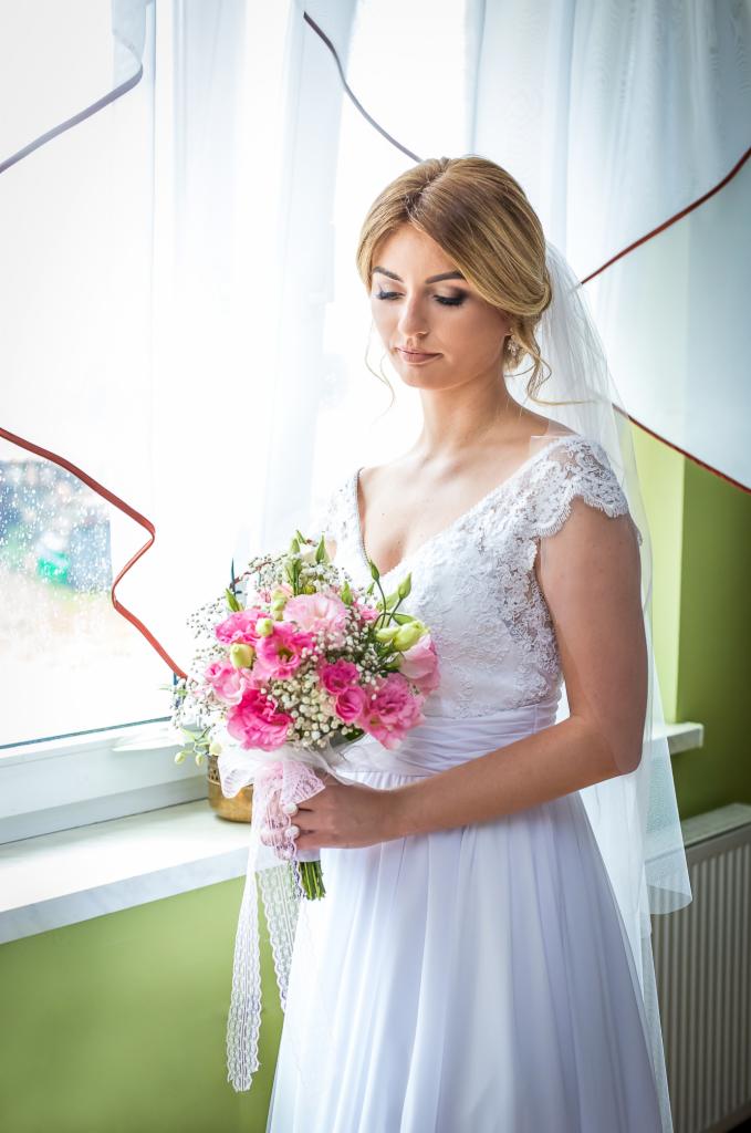 Panna młoda w białej sukni z koronkowymi rękawami, trzymająca bukiet różowych róż i gipsówki, z welonem, stojąca przy oknie.