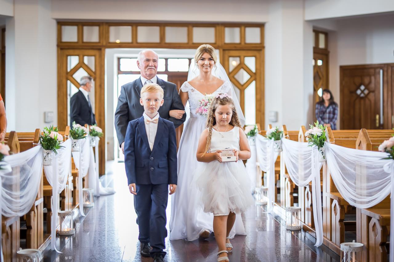 Panna młoda prowadzona do ołtarza przez ojca i dzieci niosące poduszki na obrączki, kościół z dekoracjami kwiatowymi i białymi materiałami.