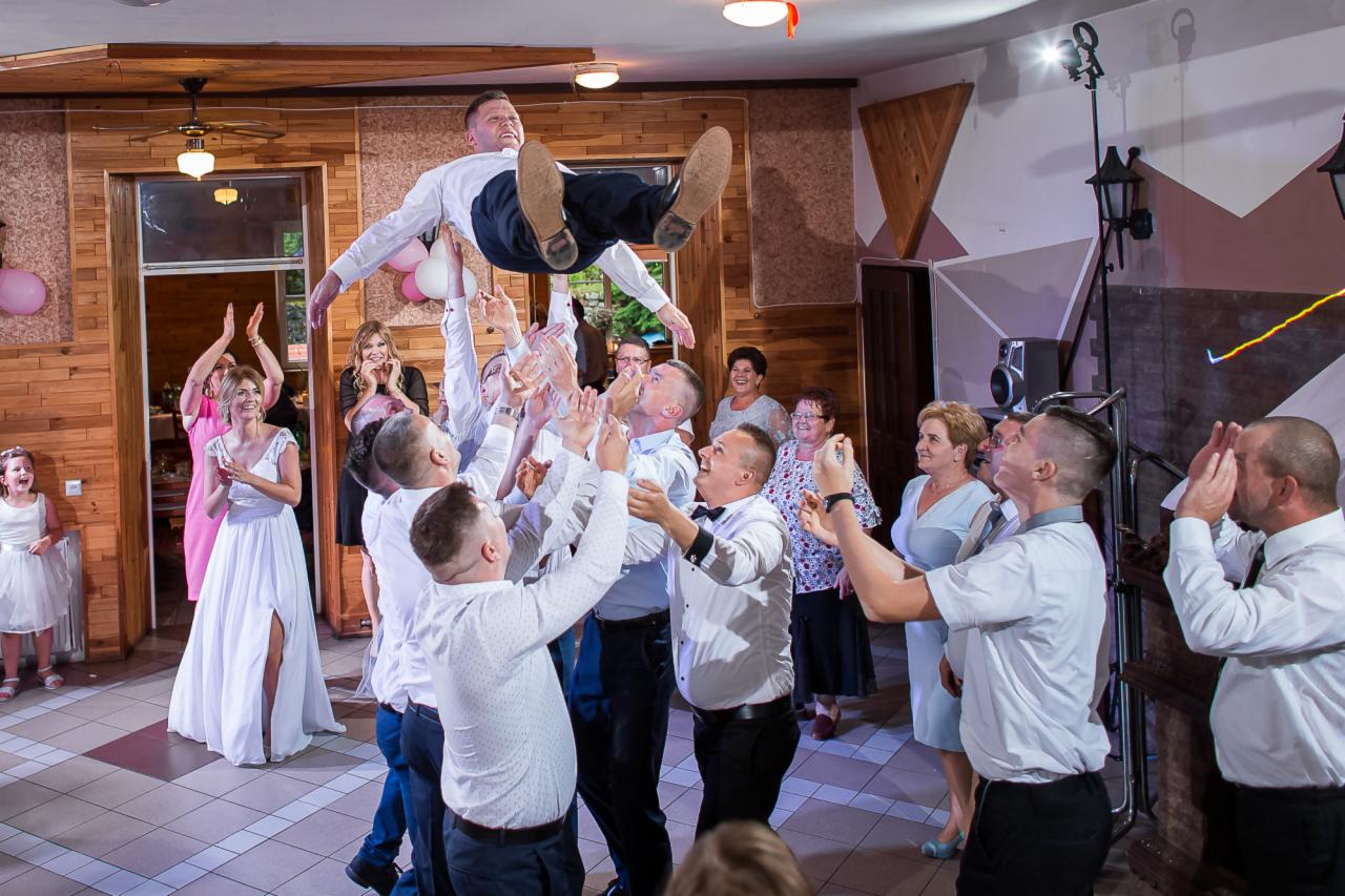 Pan młody podrzucany do góry przez gości weselnych w sali z drewnianymi elementami dekoracyjnymi, wśród uśmiechniętych kobiet w sukniach wieczorowych i dziewczynki w białej sukience.