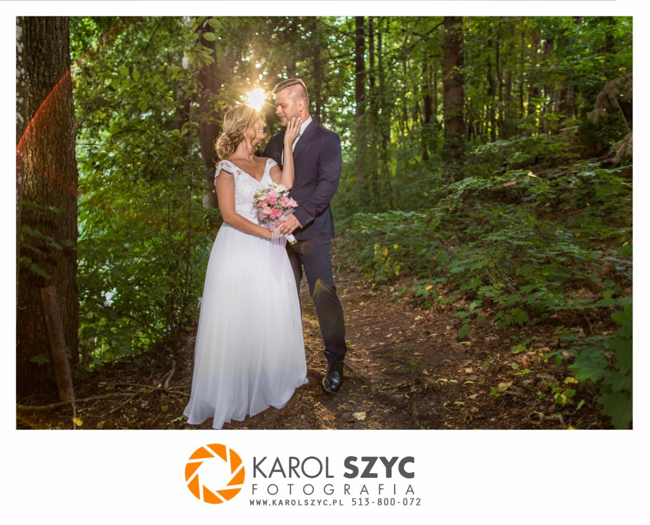 Para młoda w lesie, panna młoda w białej sukni z bukietem, pan młody w granatowym garniturze, oboje patrzą na siebie, promienie słońca przebijają przez drzewa.