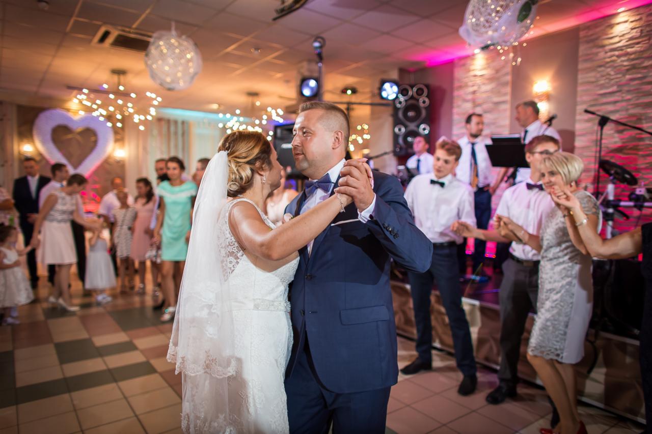Para młoda tańczy pierwszy taniec na tle zespołu muzycznego i gości weselnych, sala udekorowana lampkami i sercem.
