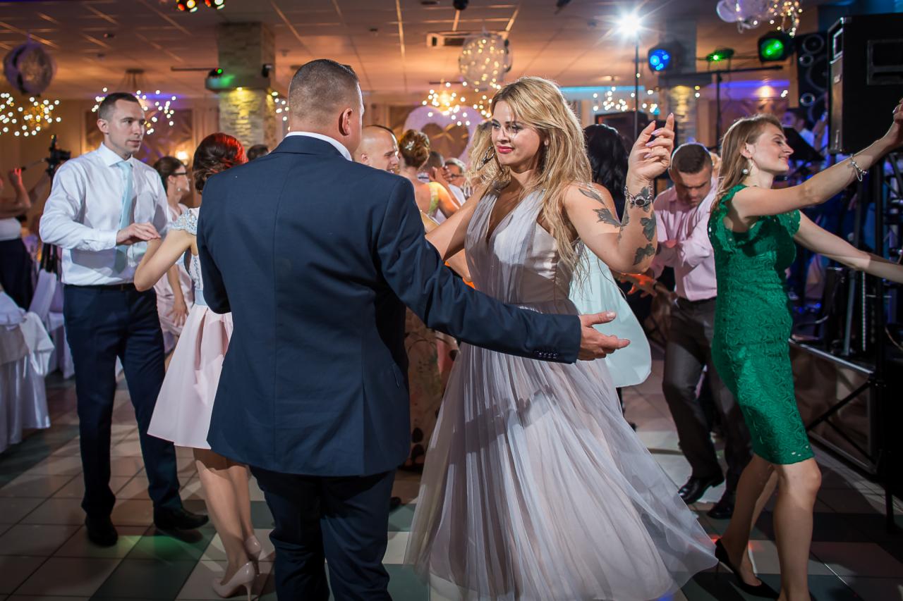 Para w tańcu na parkiecie podczas wesela, kobieta w zwiewnej sukience z tatuażami na ramieniu, mężczyzna w granatowym garniturze, w tle inni goście i dekoracje świetlne.