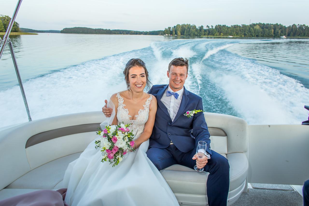 Uśmiechnięta para młoda w strojach ślubnych siedzi na białej łodzi motorowej, w tle jezioro i ślad piany po silniku.