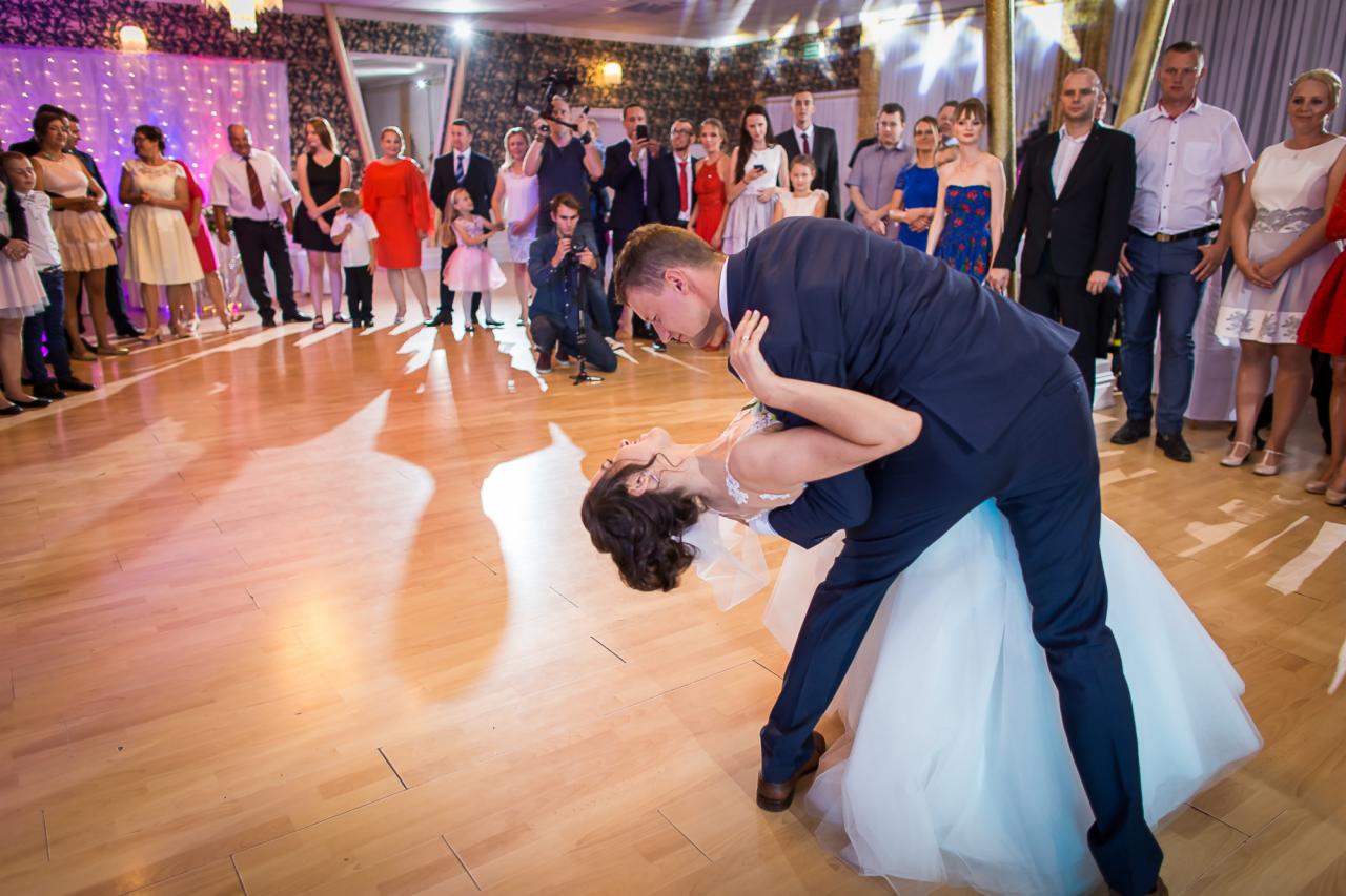 Para młoda w tańcu podczas wesela, pochylona panna młoda w białej sukni, goście weselni w tle, fotografowie robiący zdjęcia.