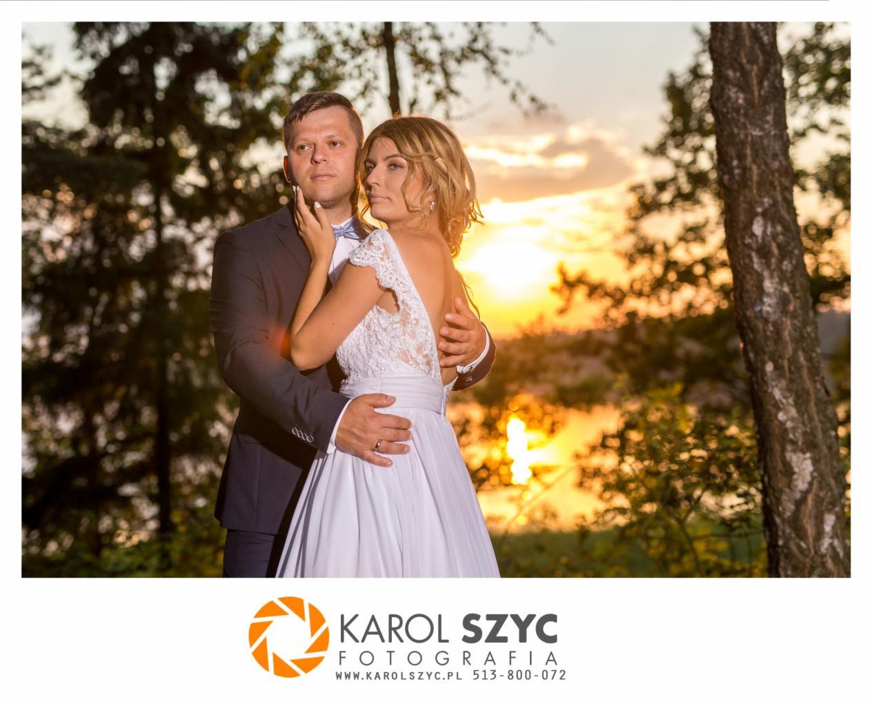 Para młoda, panna młoda w sukni z koronkowymi plecami i pan młody w granatowym garniturze, obejmujący ją na tle zachodzącego słońca nad jeziorem, widoczne drzewa.