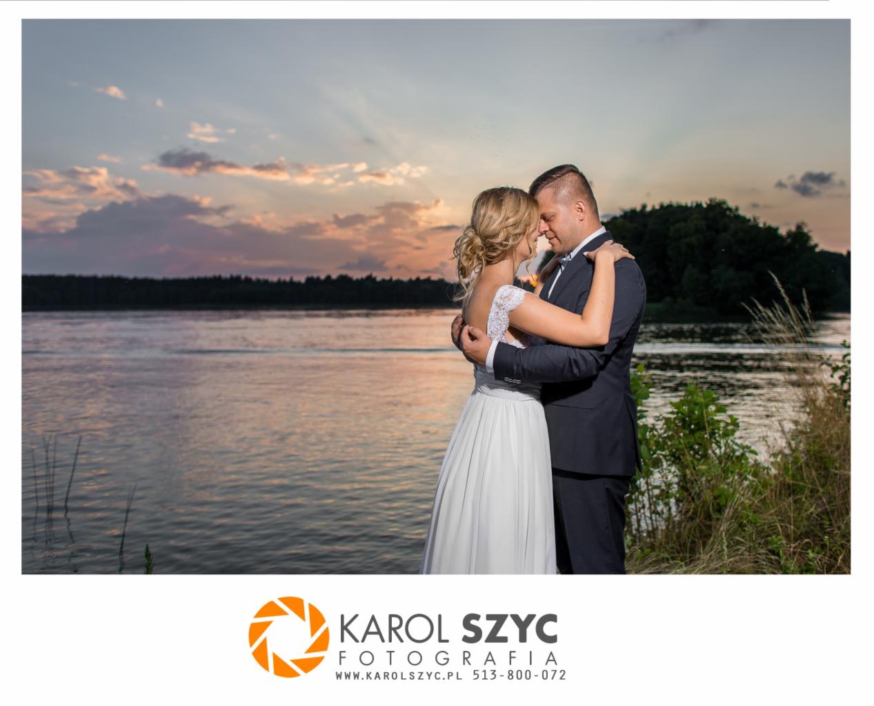 Para młoda w objęciach na tle jeziora o zachodzie słońca, panna młoda w sukni z koronkową górą, pan młody w ciemnym garniturze z muszką.