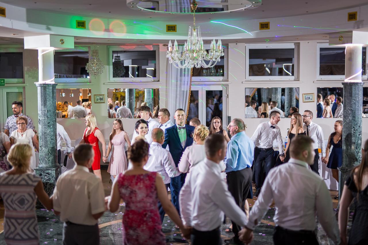 Para Młoda w otoczeniu gości weselnych tańczących w kole, oświetlonych dekoracyjnym żyrandolem i kolorowymi światłami na sali balowej.