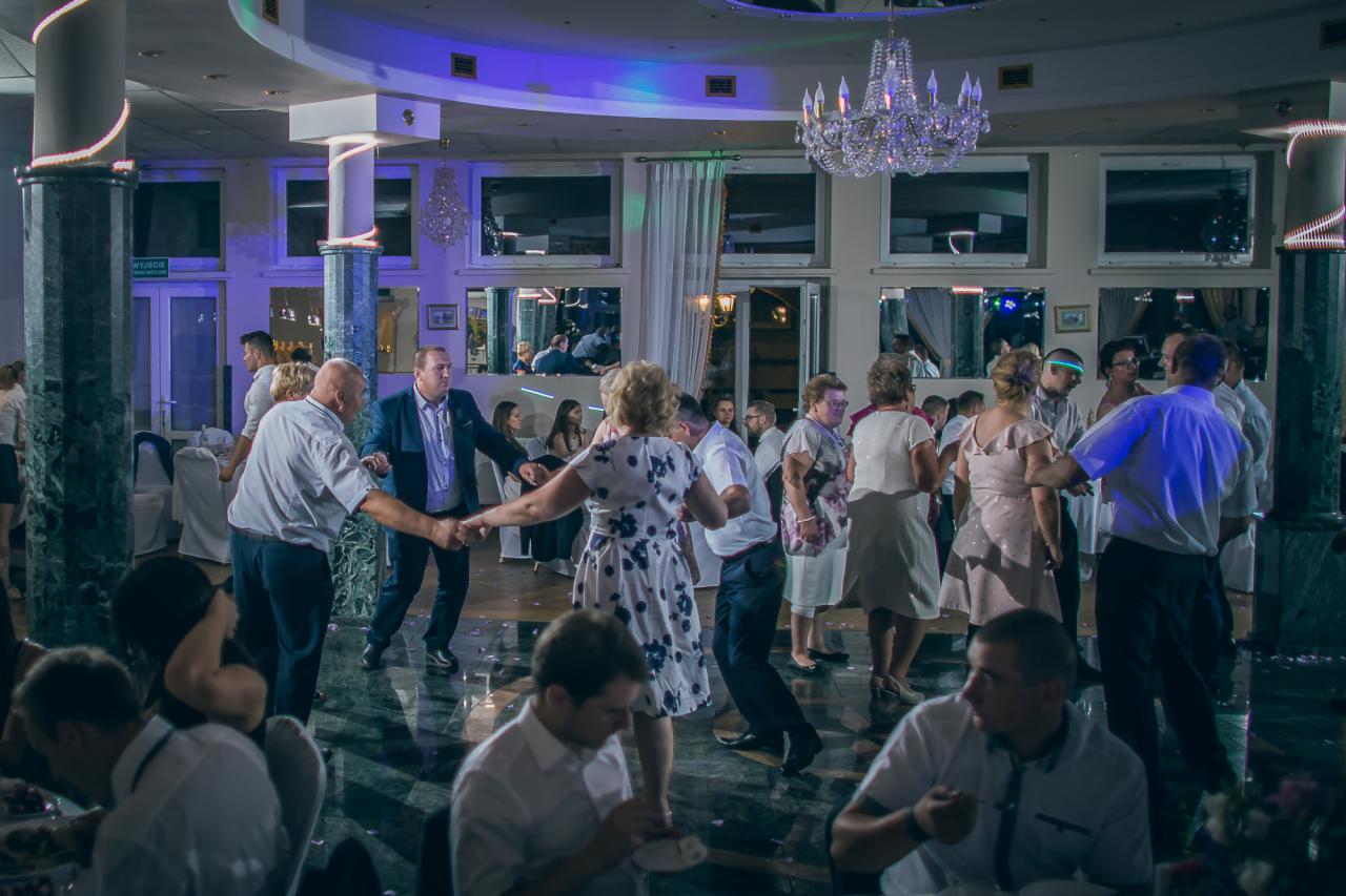 Goście weselni tańczą w sali balowej z marmurową podłogą, pod kryształowym żyrandolem, w tle okna z lustrami.