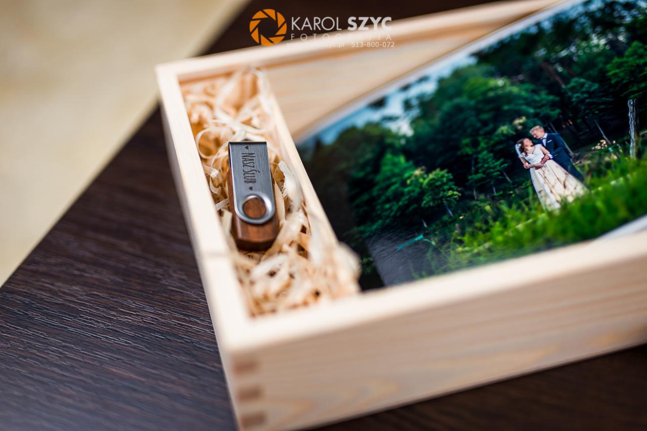 Drewniane pudełko z fotografią ślubną pary w plenerze i pendrivem z napisem 'Nasz Ślub' w wypełnieniu z rafii.