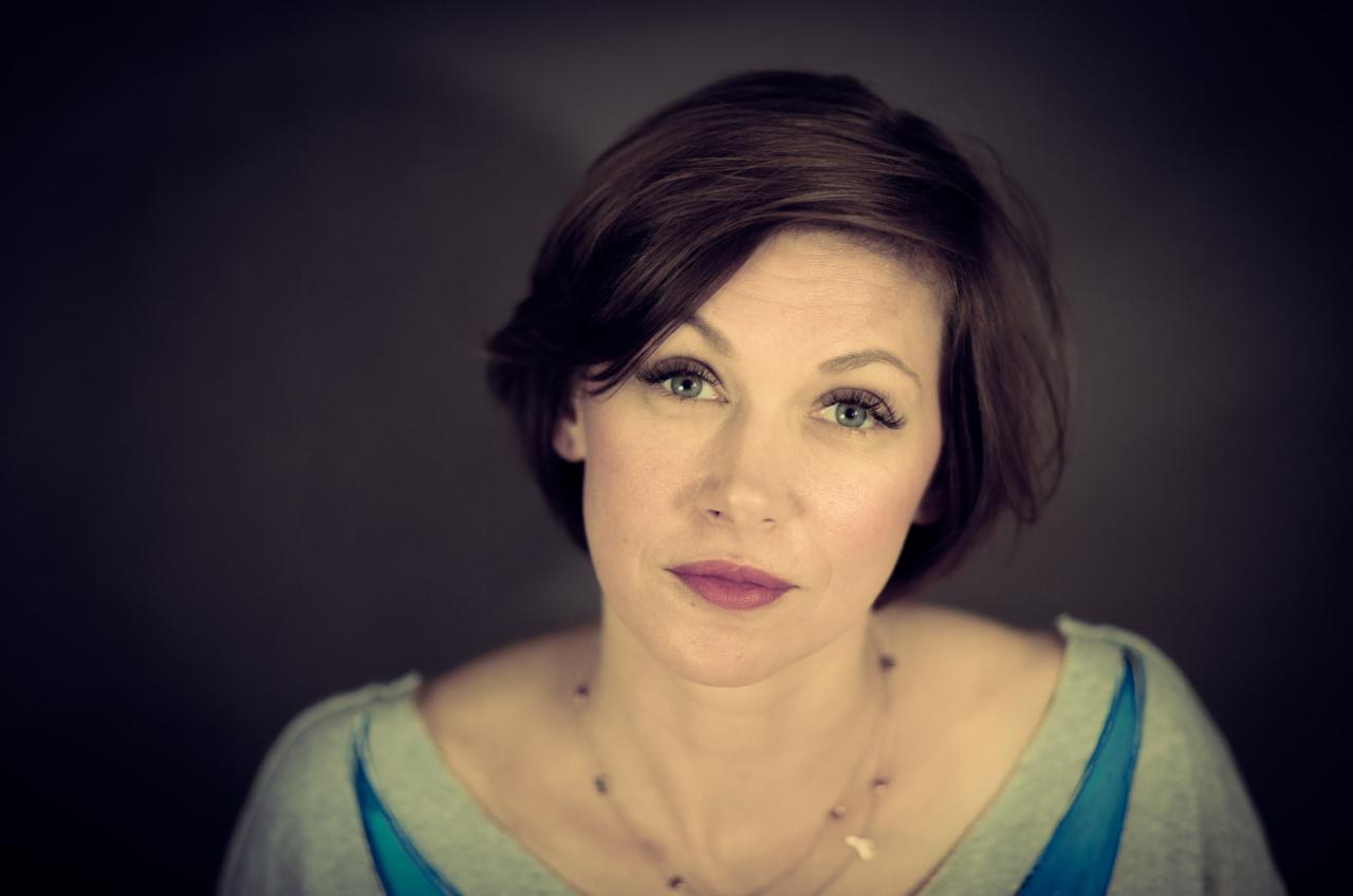 Portret kobiety z krótkimi brązowymi włosami, zielonymi oczami i delikatnym makijażem na ciemnoszarym tle, ubrana w szarą bluzkę z niebieskimi akcentami i subtelną biżuterię.