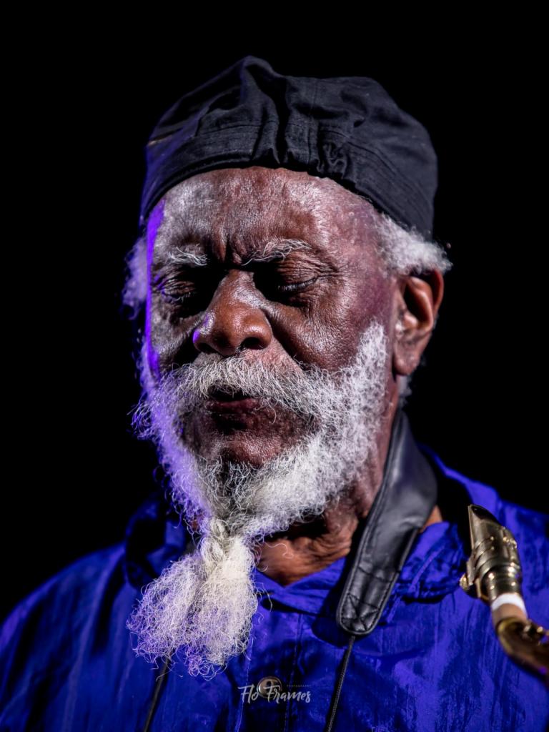 Portret starszego muzyka jazzowego z zamkniętymi oczami w czarnej czapce i z siwą brodą zaplecioną w warkocz, trzymającego saksofon na szyi w niebieskiej kurtce na czarnym tle.