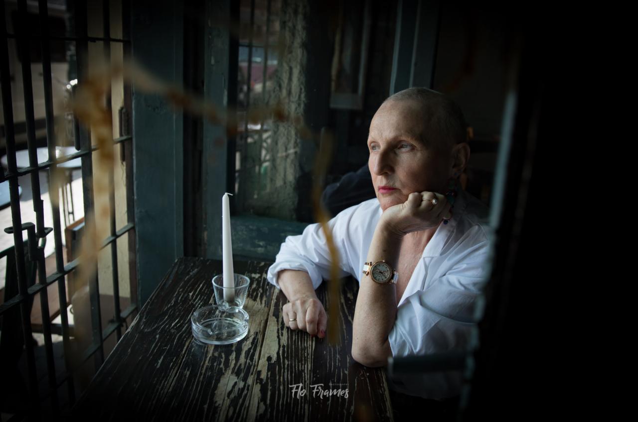 Portret kobiety w białej koszuli, siedzącej przy drewnianym stole z popielniczką i świecą, zamyślona twarz, zegarek na ręce, w tle okno z kratami.