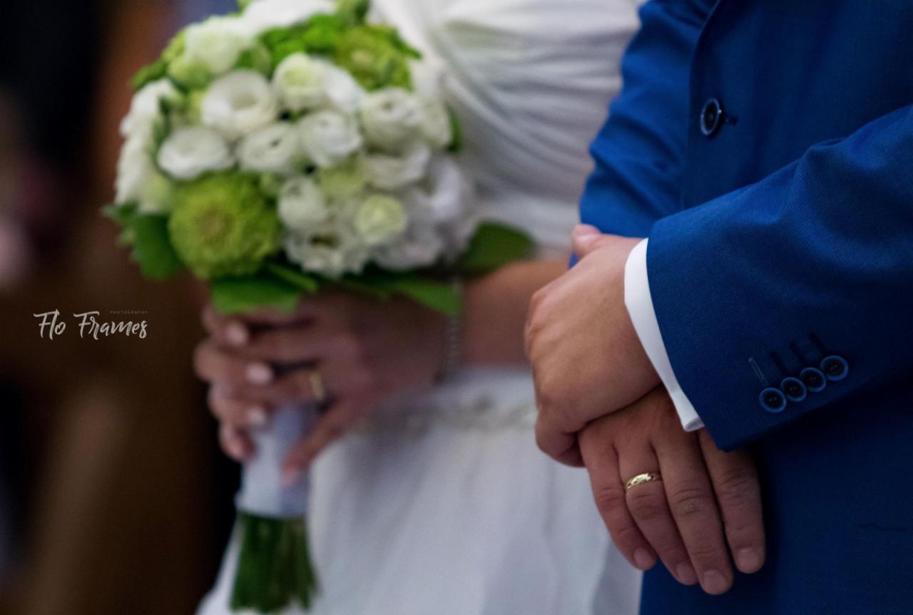 Dłonie pary młodej, on w błękitnym garniturze, ona trzyma bukiet białych i zielonych kwiatów, widoczne obrączki.