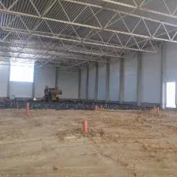 Wnętrze budowanej hali z widoczną konstrukcją stalową dachu, betonowymi słupami, ścianami z płyt warstwowych, przygotowanym podłożem z kamienia i pomarańczowymi rurami instalacyjnymi w ziemi...