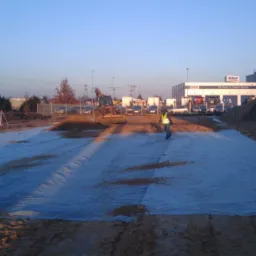 Przygotowanie podłoża pod drogę: rozłożona geowłóknina, pracownik w kamizelce odblaskowej, spychacz i koparka w tle, budynek DAF w oddali.