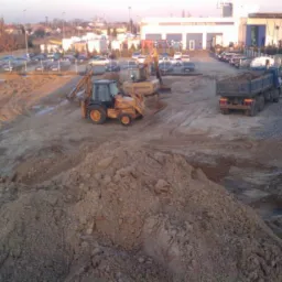 Żółta koparko-ładowarka i wywrotka na placu budowy, widoczne sterty ziemi i kruszywa, w tle budynek firmy transportowej i zaparkowane samochody osobowe.