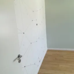 Ściana z geometrycznym wzorem w pokoju, świeżo położone panele podłogowe, widoczna część drzwi z klamką i fragment ściany pomalowanej na szaro.
