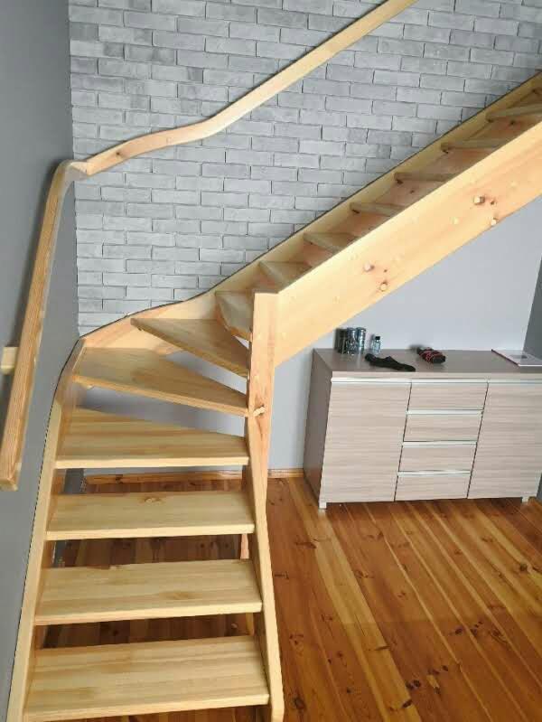 Drewniane schody spiralne z jasnego drewna, prowadzące na antresolę, z widoczną balustradą i fragmentem ściany z imitacją cegły.