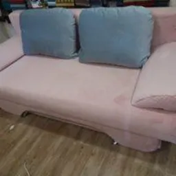 Różowa sofa z dwoma niebieskimi poduszkami na drewnianej podłodze, widoczna część metalowej nogi.