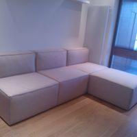 Jasnoszara, modułowa sofa narożna z widocznym przeszyciem na krawędziach, ustawiona na drewnianej podłodze w jasnym pomieszczeniu.