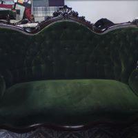 Antyczny, zielony welurowy fotel z ciemnobrązowymi, rzeźbionymi elementami dekoracyjnymi. Widoczny fragment ściany w tle.