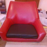 Czerwony fotel po renowacji tapicerskiej z brązowym siedziskiem, widoczne niedoskonałości oświetlenia i tło z materiałami.