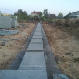 Wykop z betonową wylewką i ułożonymi bloczkami fundamentowymi wzdłuż wykopu, widoczne bloczki czekające na ułożenie oraz domy w tle.