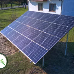 Panel fotowoltaiczny zamontowany na metalowej konstrukcji na trawniku przed domem jednorodzinnym, widoczne odbicie słońca na panelach.