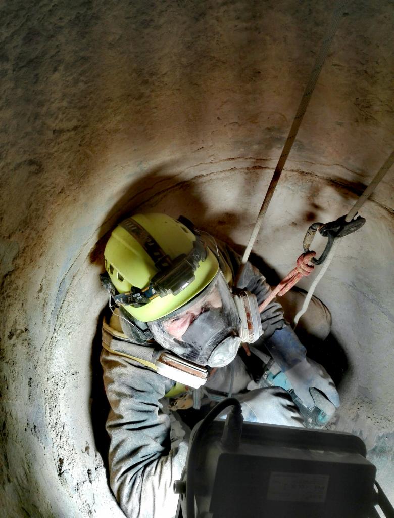 Pracownik w kasku i masce ochronnej, zabezpieczony linami, pracuje wewnątrz betonowego szybu, oświetlony lampą.