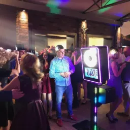 Uczestnicy imprezy w strojach wieczorowych korzystają z fotobudki z ekranem dotykowym i oświetleniem LED na drewnianym parkiecie, w tle ceglany filar i dekoracje sufitowe.