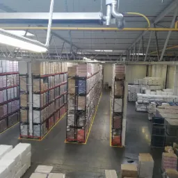 Wnętrze magazynu z wysokimi regałami wypełnionymi zapakowanymi towarami, widok z g&oacute;ry, oświetlenie jarzeniowe, ż&oacute;łte oznaczenia na podłodze.