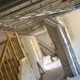 Montaż metalowej konstrukcji pod sufitem podwieszanym w trakcie remontu, widoczne przewody elektryczne i wentylacyjne, surowe ściany i schody drewniane.