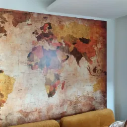 Ściana z tapetą przedstawiającą mapę świata w stylu vintage, widoczny fragment sofy w kolorze musztardowym i zasłony w podobnym odcieniu.