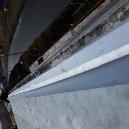 Szara rynna dachowa z widocznymi kroplami deszczu, w tle ulica i zieleń, ujęcie z góry.