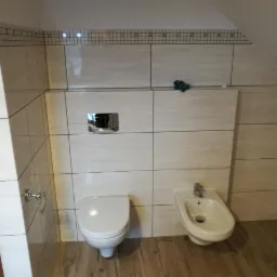 Wykończona łazienka na poddaszu z beżowymi płytkami, wiszącą miską WC i bidetem, chromowanymi elementami armatury i drewnopodobną podłogą.