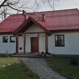 Nowoczesny dom jednorodzinny z jasną elewacją, czerwoną dachówką i drewnianym gankiem, otoczony trawnikiem i ścieżką z drobnego żwiru.