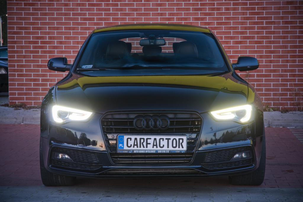 Czarny samochód marki Audi z włączonymi światłami, tablicą rejestracyjną z nazwą firmy 'Carfactor' na tle ceglanej ściany.