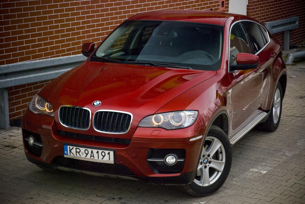 Czerwone BMW X6 zaparkowane na brukowanej nawierzchni z ceglanym murem w tle, widoczne tablice rejestracyjne KR 9A191.
