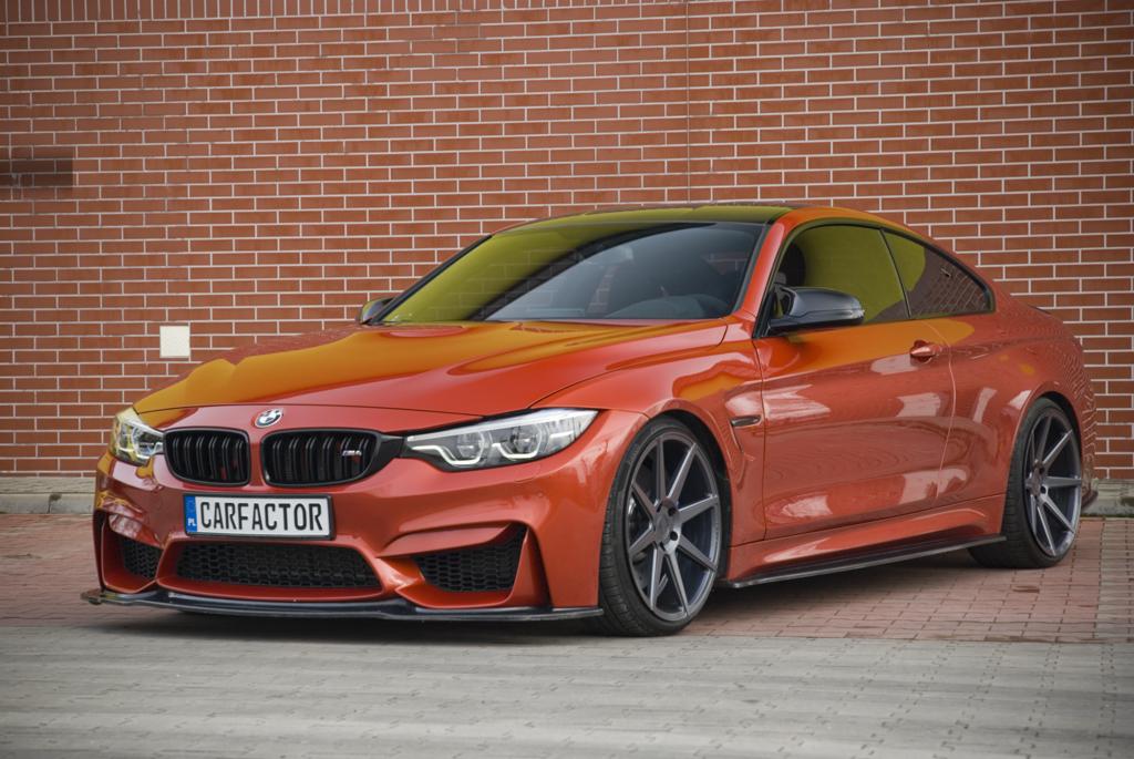 Pomarańczowe sportowe coupe z czarnym dachem na tle ceglanej ściany i szarej kostki brukowej. Widoczna tablica rejestracyjna z nazwą 'CARFACTOR'.