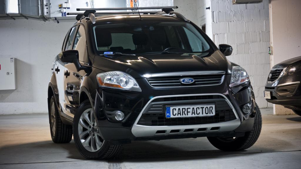 Czarny Ford Kuga z relingami dachowymi, zaparkowany w garażu podziemnym, widoczna tablica rejestracyjna z nazwą CARFACTOR.