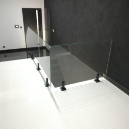 F.P.H.U. EK-GLASS Beata Kocoń - Nowoczesna balustrada ze szkła hartowanego, zamocowana na czarnych wspornikach, oddziela schody w minimalistycznym wnętrzu z białymi płytkami i ciemną ścianą.