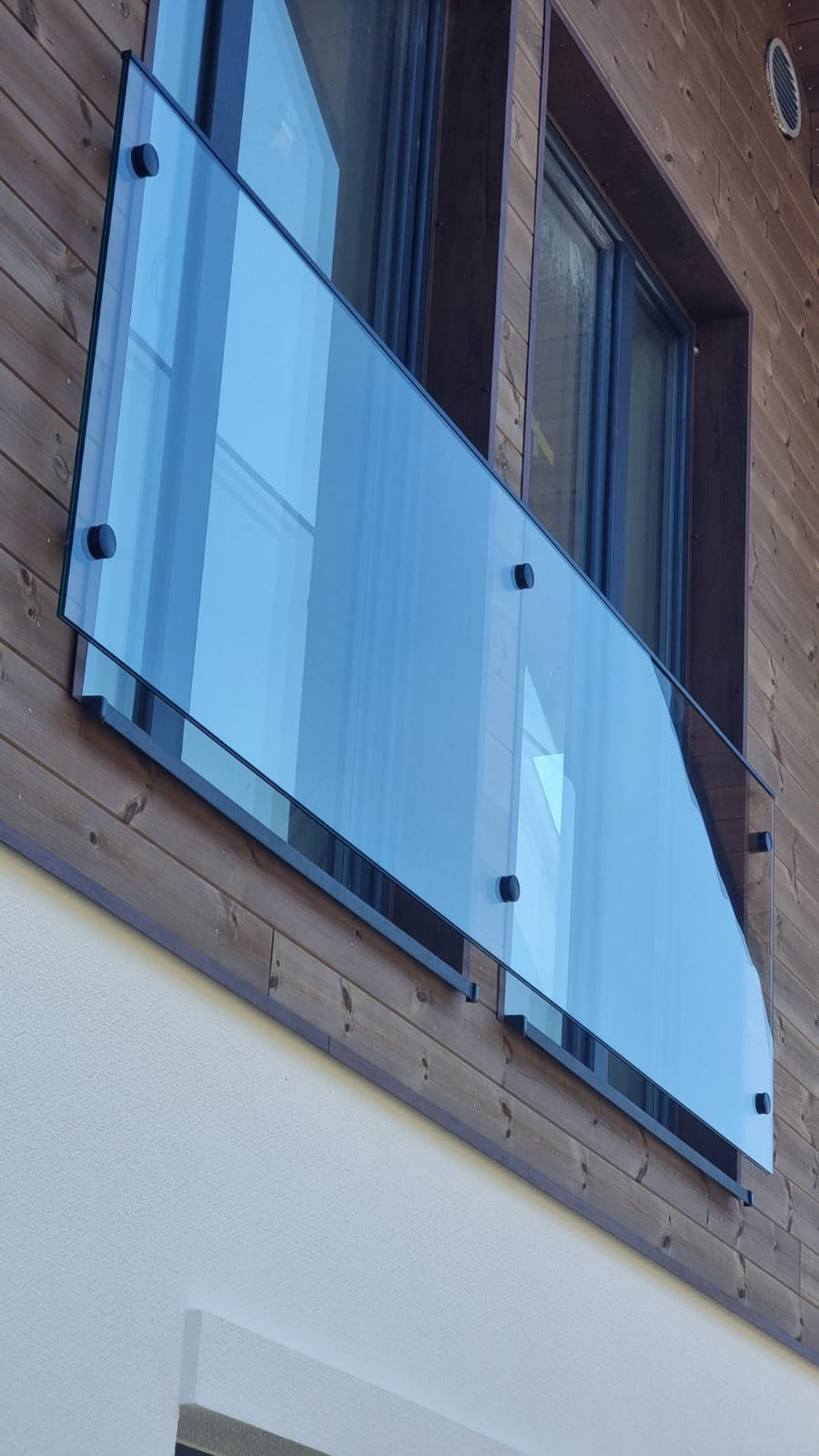 Szklana balustrada balkonowa z mocowaniami punktowymi na tle drewnianej elewacji. Nowoczesny design, czyste linie i przezroczystość szkła.